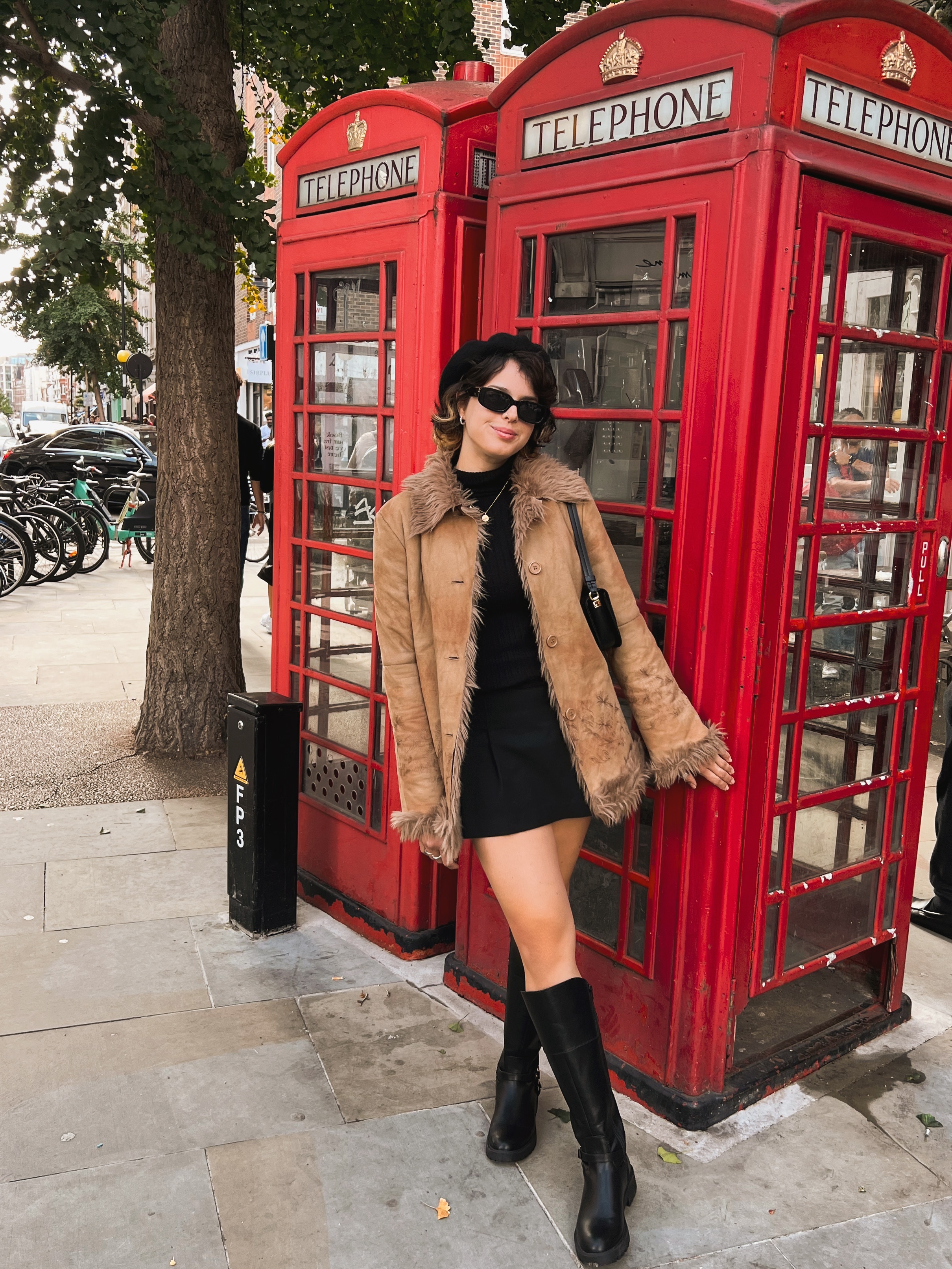 A young woman in short black dress, black boots, and faux fur coat with sunglasses poses in front of a red phone booth in London